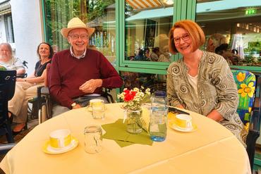 Auf diesem Bild ist ein Heimbewohner mit seiner Tochter abgebildet. Beide sitzen an einem Tisch und lächeln in die Kamera.