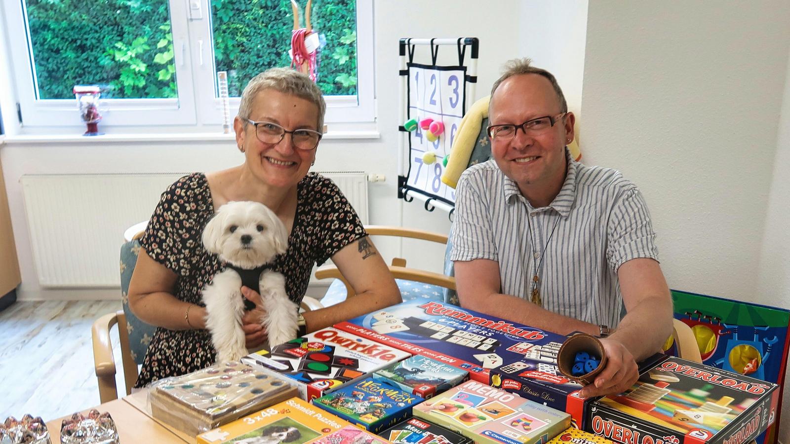 Auf diesem Bild ist Frau wirth mit ihrem Hund zu sehen. Danneben sitzt Herr Wolf-Kather unser Leiter der Mitgliederbetreuung. Im Vordergrund des Bildes befinden sich diverse Tischspiele.