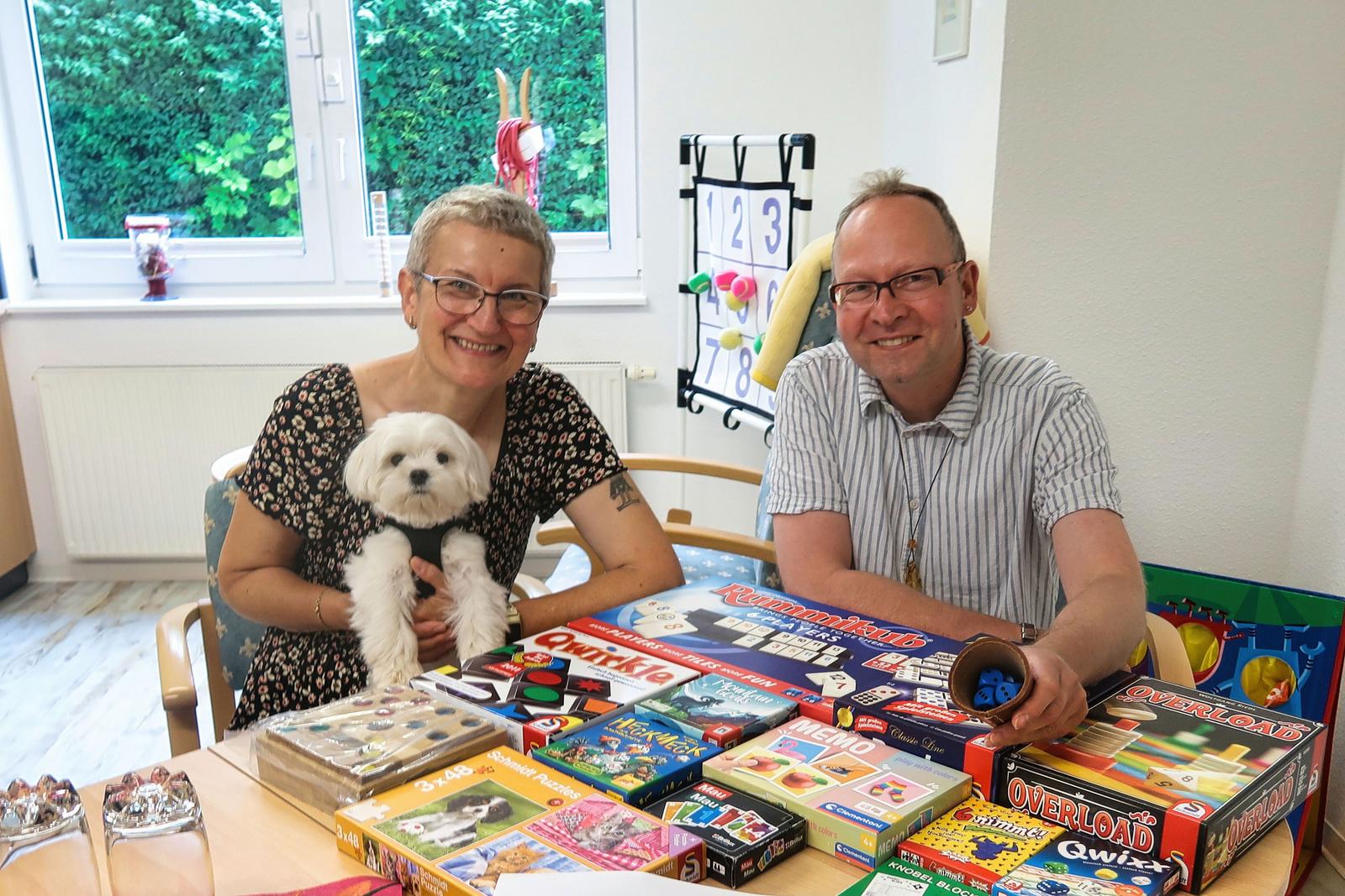 Auf diesem Bild ist Frau wirth mit ihrem Hund zu sehen. Danneben sitzt Herr Wolf-Kather unser Leiter der Mitgliederbetreuung. Im Vordergrund des Bildes befinden sich diverse Tischspiele.