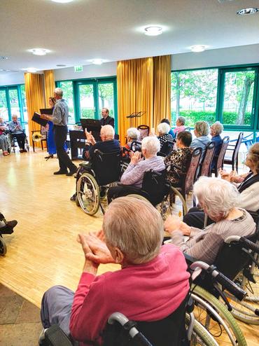 Auf diesem Bild sind eine vielzahl älterer Menschen aus dem Pflegeheim zu sehen. Die Perspektive bildet diese Menschen von Hinten ab. Insgesamt wirkt das Bild recht dynamisch.