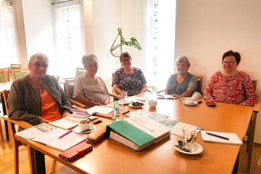 Auf diesem Bild ist die Leitung unseres Seniorenchors zu sehen. Sie sitzen am Tisch uns schauen freudig in die Kamera.