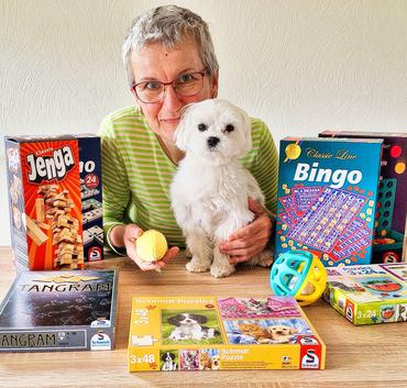 Auf dem Bild ist Frau Annett Wirth, unsere Spieleexpertin zu sehen. Sie lächelt in der Kamera und posiert zusammen mit einigen Spielen und ihrem Hund. 