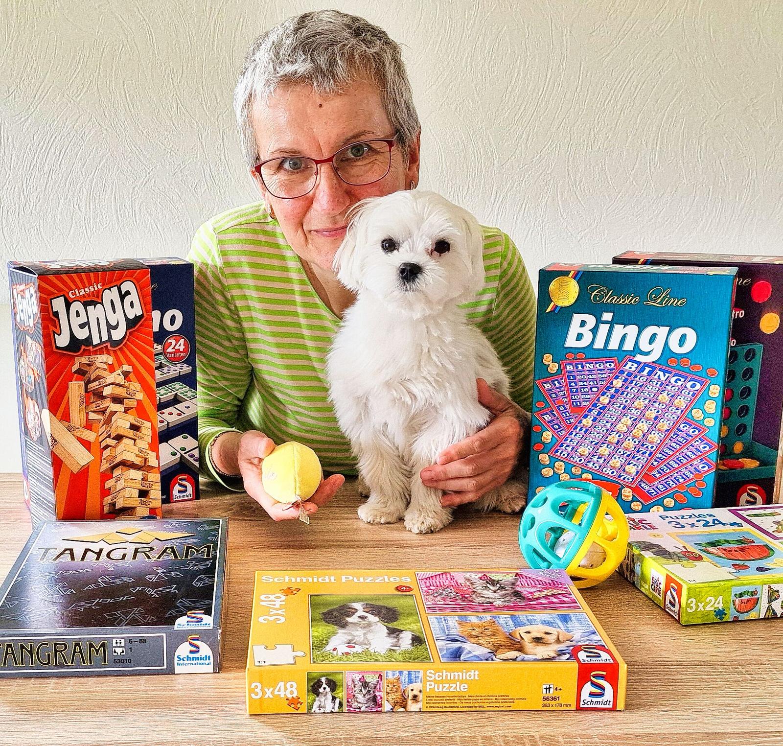 Auf dem Bild ist Frau Annett Wirth, unsere Spieleexpertin zu sehen. Sie lächelt in der Kamera und posiert zusammen mit einigen Spielen und ihrem Hund. 
