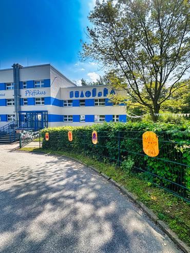 Auf diesem Bild sieht man unsere Kindertagsstätte Pfiffikus von außen. Am Zaun sind die kleinen Kunstwerke der Kinder angebracht. Das Wetter ist sehr freundlich.