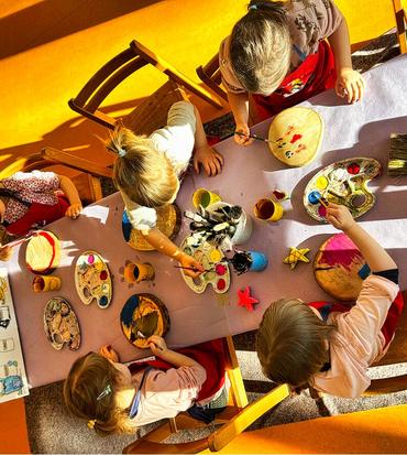 Auf diesem Bild sind die Kinder von oben fortografiert. Sie sitzen an einem Tisch und bearbeiten die Kunstwerke für den Zaun. Es wird gemalt, gebastelt und gepinselt. 