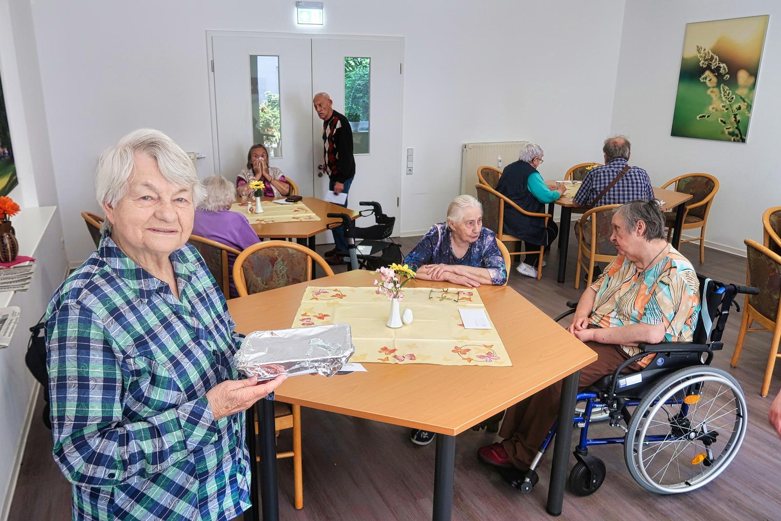 Auf diesem Bild sieht man viele unserer Bewohner am gemeinsamen Mittagstisch. Frau herberger steht im Vordergrund und lächelt mit ihrer Mahlzeit in der Hand in die Kamera. Die Atmosphäre ist gesellig und entspannt.