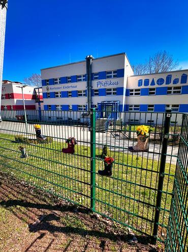 Auf diesem Bild sieht man unsere Kindertagsstätte Pfiffikus von außen. Am Zaun sind die kleinen Kunstwerke der Kinder angebracht. Das Wetter ist sehr freundlich.