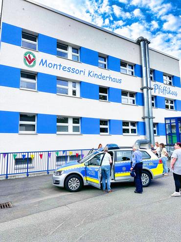Auf diesem Foto ist unsere Kita Pfiffikus zu sehen. Vor dem Haus steht ein Polizeiauto. Davor stehen ein paar Polizisten die sich mit den Erziehern nett unterhalten.